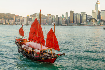 Traditional red Chinese Junk boat sailing in front of Hong Kong skyline during the day. Boat has three red sails.