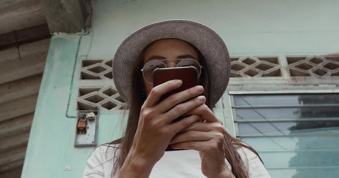 Young woman in stylish hat browsing modern smartphone while standing near weathered building