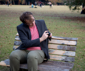 A man sat on a park bench taking pictures with his digital camera
