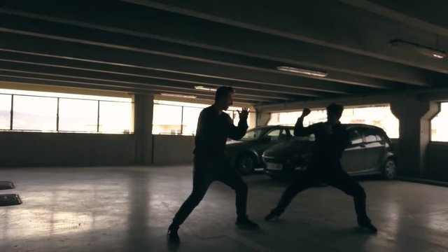 Wide view of men fighting in front of cars in a parking garage. Leaping over a man and spinning. Actors fight in a movie scene. Stunt men fighting on a movie set.