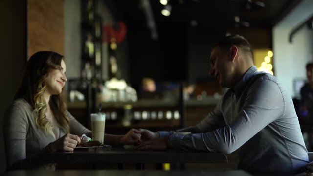 Beautiful couple/lovers sitting in a cafe touching their hands, looking at, talking to each other, smiling together. There is waitress working background. Woman with big boobs/breast very happy
