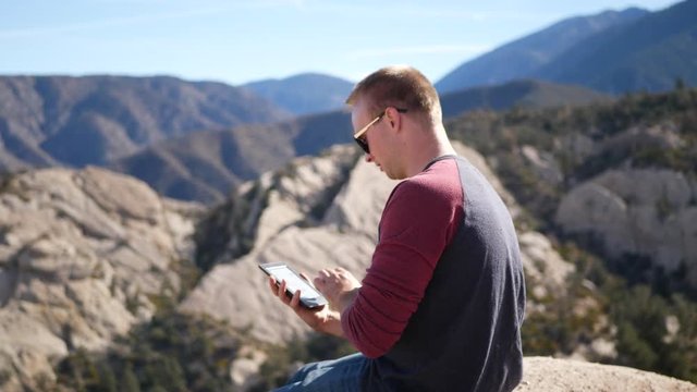 A man flipping through pages as he reads a book on his e reader tablet device on a nature camping trip adventure in the mountains.