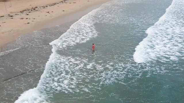 Aerial, zoom in, drone shot of a woman walking in the water, while waves hit the paradise beach, near Trincomalee city, on a cloudy day, in Gokanna, in the Eastern Province of Sri Lanka, Asia
