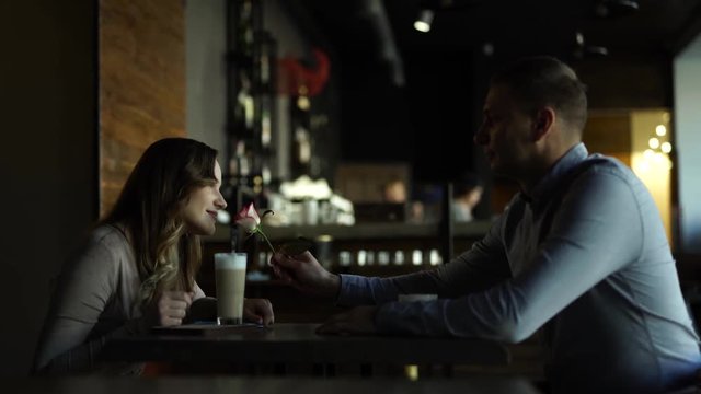 Couple/lovers sitting in a cozy cafe looking at each other. Handsome man giving a woman in decolette dress/big breast a white-red rose. Blonde is smelling a flower and smiling fully. Love in air