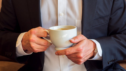 Man holding a cup of coffee with cream in white ceramic cup, dressed in white dress shirt and black business suit and wearing a watch