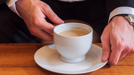 Man sitting at a table drinking coffee with cream in a white ceramic cup on a small white ceramic plate, wearing white dress shirt, black business suit and a watch