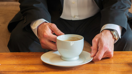 Man sitting at a table drinking coffee with cream in a white ceramic cup on a small white ceramic plate, wearing white dress shirt, black business suit and a watch