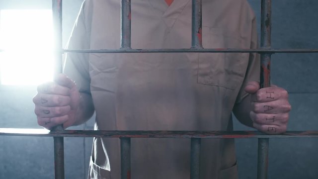 Medium shot of a prisoner, with "love" and "hate" tattooed across his knuckles, clutches the bars of his prison cell as the camera dollies in, then he walks away.