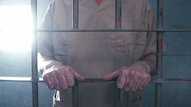Medium shot of a prisoner with "love" and "hate" tattooed across his knuckles as the camera dollies in, then he walks away.