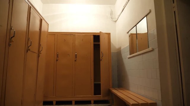 Walking away from an open locker door in a narrow empty school change room with creepy orange lighting and numbered wooden doors