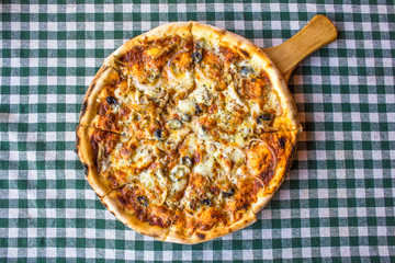 whole round pizza on a wooden plate with a handle on a table with a checkered tablecloth. hot pizza top view
