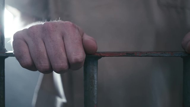 Close-up tracking shot from left to right of a prisoner's hands holding onto the bars of his cell door. When the camera stops moving he walks off screen.