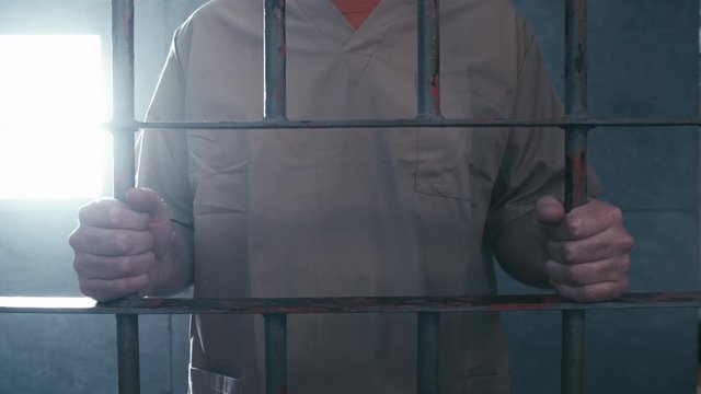Medium dolly shot of a prisoner holding on to the bars of his cell door. When the camera stops moving, he walks off screen.