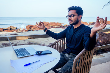 handsome and successful indian man in a stylish well-dressed freelancer working laptop meditating in the lotus position on the beach.freelance and remote work.businessman india namaste and om ocean - obrazy, fototapety, plakaty