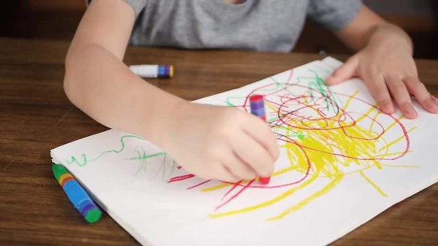 Close-up of a small child drawing bright color crayons or pencils on paper while sitting at a wooden table. The development of children of preschool age, creativity.