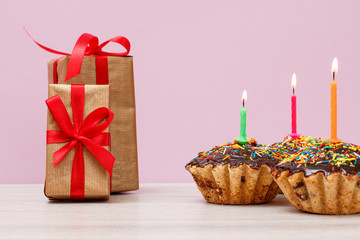 Gift boxes and birthday cupcakes with burning festive candles on lilac background.