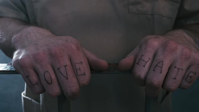 Extreme close-up shot of a prisoner's hands with "love" and "hate" tattooed across his fingers as he clutches the bars of his prison cell while the camera does a parallax move.