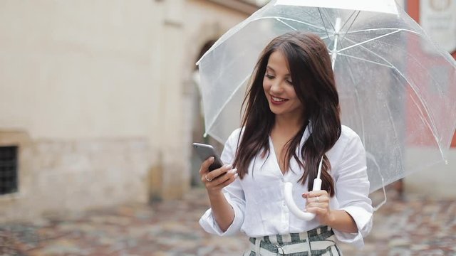 Beautiful young business woman using smartphone walking on the street in rainy weather, smiling, holding umbrella. Communication concept