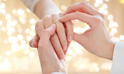 gay, homosexuality, same-sex marriage and lgbt concept - close up of lesbian couple hands putting on wedding ring over festive lights background