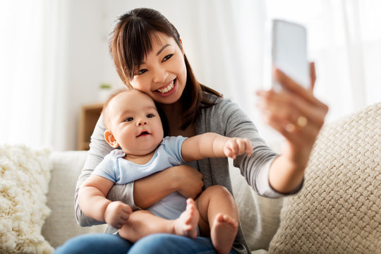 family, technology and motherhood concept - happy young asian mother with little baby son taking selfie by smartphone at home