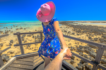 Woman in hat holding hand of her friend on wooden path at Stromatolites Hamelin Pool. Marine Reserve in Shark Bay, Western Australia. A girl enjoys of Australian travel destination. Follow me POV.