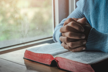 Close up hands of young male praying on holy Bible at home.