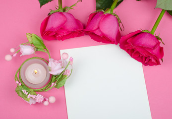Beautiful pink roses and a burning candle on a blank white greeting card against a bright pink background (flat lay, top view)