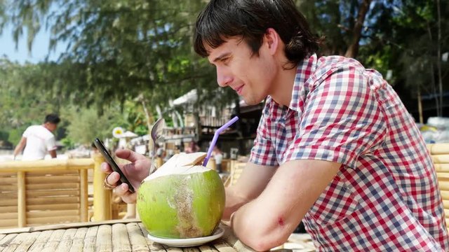 Handsome young businessman in sunglasses drink fresh coconut juice in a beach cafe with sea view on palm trees on background and uses mobile phone. 3840x2160