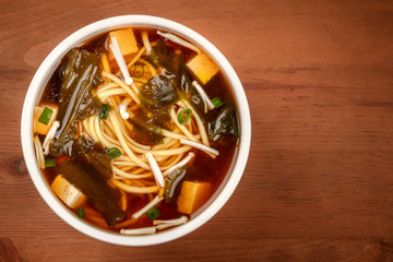 Miso shiru soup with tofu, scallions, noodles, wakame seaweed and enoki mushrooms on a rustic wooden background with copy space