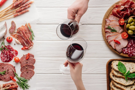 partial view of couple clinking glasses with red wine at white tabletop with meat appetizers - Powered by Adobe
