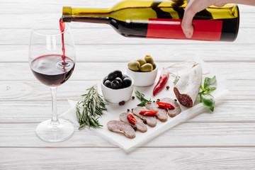 partial view of woman pouring red wine into glass on white wooden tabletop with meat appetizers near by