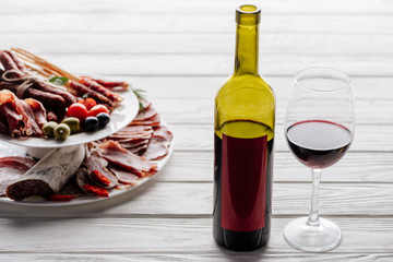 close up view of bottle and glass of red wine and various meat appetisers with olives on white wooden tabletop