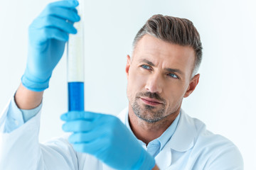 selective focus of handsome scientist looking at test tube with blue reagent isolated on white