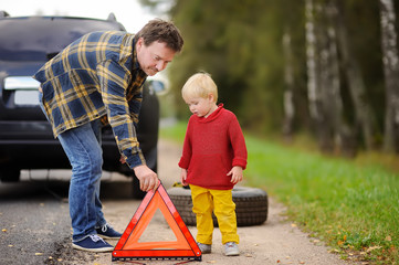 Father and his little son repairing car and changing wheel together on summer day.
