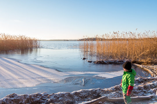 Young child in winter clothing looking at a beautiful cold sunny scenic tranquil winter landscape of ice, water and reed against a clear blue sky.