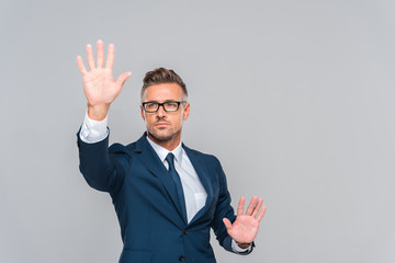 handsome businessman in blue suit and glasses touching something isolated on grey, artificial...