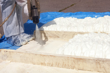 Technician dressed in a protective white uniform spraying foam insulation using Plural Component Spray Gun. Spraying polyurethane foam for roof and energy saving - obrazy, fototapety, plakaty