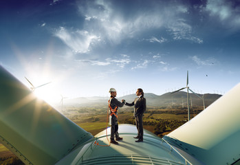 Happy engineer and businessman shake hands after good work. They standing a top of windmill. Around...