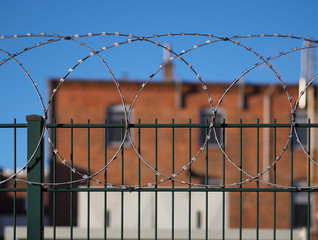 Green steel fence with barbed wire on top and red brick building in the background (out of focus)