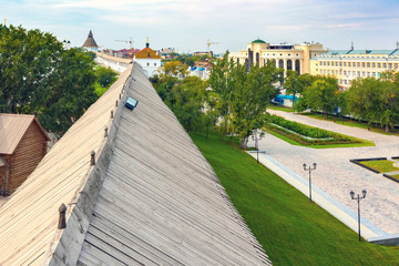 Fototapeta premium The wooden roof of the medieval white stone wall between the buildings of the city and courtyard on a sunny day with white blue clouds. Historical and architectural complex Astrakhan Kremlin, Russia.