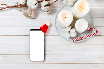 Fototapeta na wymiar Smart phone with Christmas hat on white wooden holiday background with cotton branch, candles and candy cane. Close-up, top view. Mock up. Christmas and New Year concept. Copy space