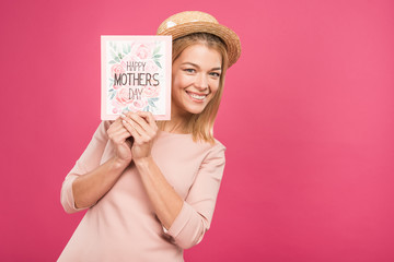 smiling woman holding "happy mothers day" greeting card, isolated on pink