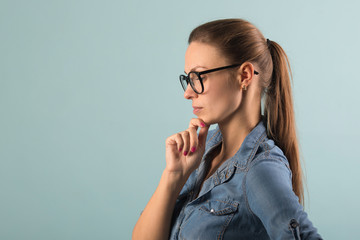 beautiful young girl in a denim shirt and glasses on a blue background with a serious thinking face