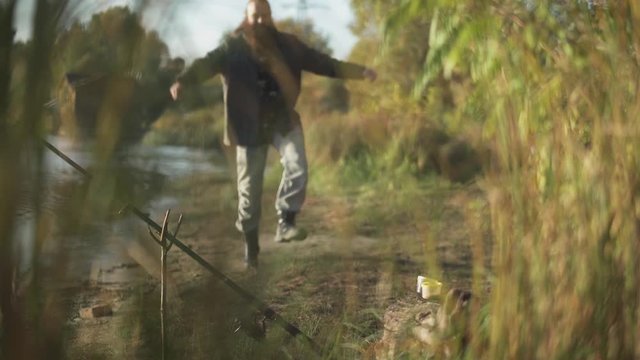 Funny bearded man moves to the camera in joking way, jumping and making funy moves. Fishing rod is fixed in special stick on the river bank