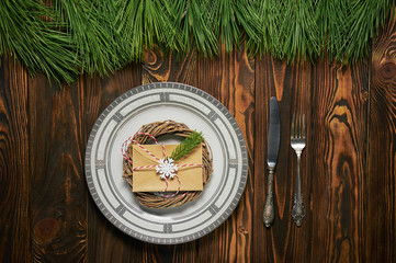 Fototapeta na wymiar christmas table setting at brown wooden background with white plate, silver cutlery and paper envelope.