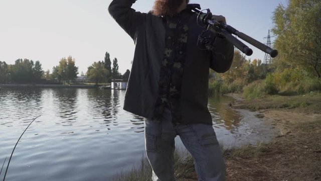 Fisherman with long beard stends on the river bank with fishing rods. The man looks into the distance, covering his eyes with his hand from the sun. Slow motion.