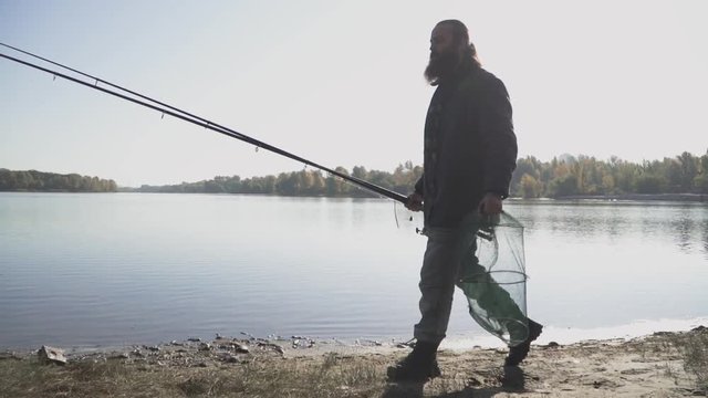 Adult fisherman with long beard walks near the river with fishing rods and ambush hoop net. Slow motion.
