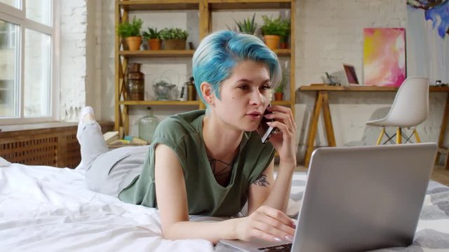 Tracking shot of cheerful young woman with blue hair lying on bed and chatting on mobile phone while typing on laptop