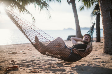 A young man is lying in a hammock at sunset by the ocean. Handsome guy is resting in a hammock...