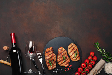 Grilled pork steaks on stone with bottle of wine, wine glass, knife and fork on rusty background